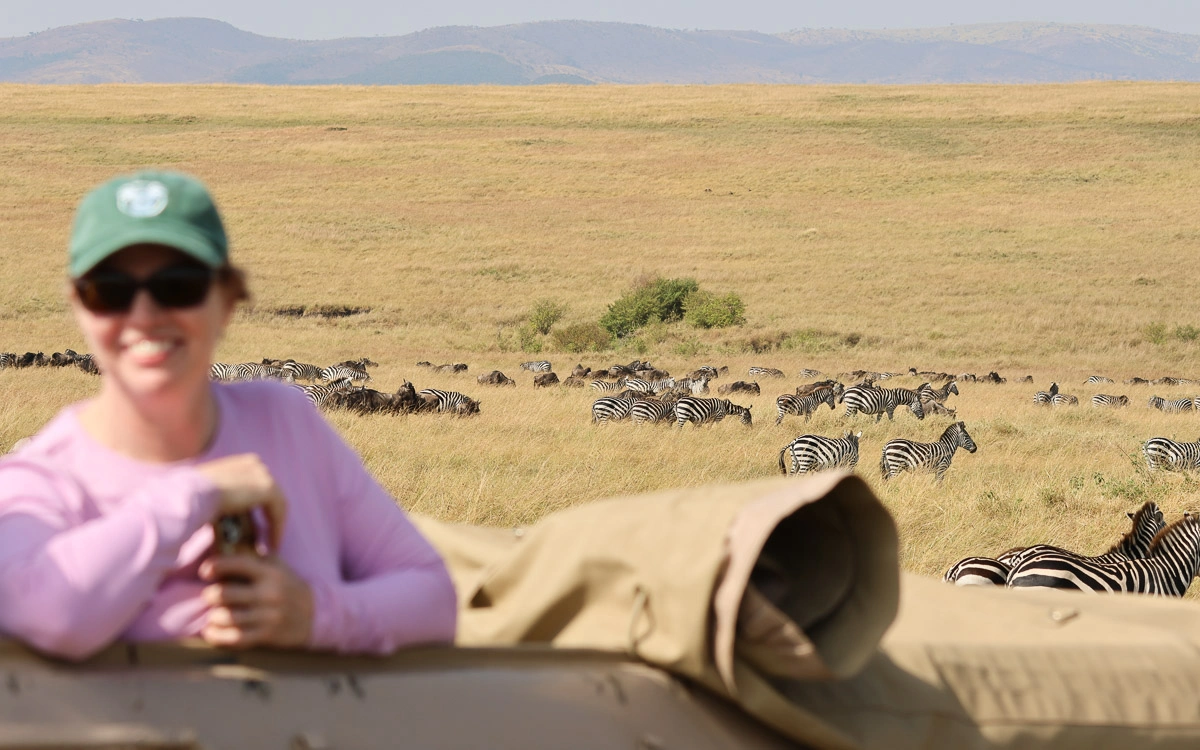 A guest smiling on a game drive as a large herd of zebra and wildebeest graze in the distance